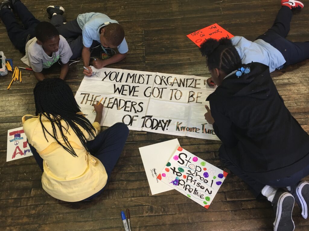 photo: a group of five Black middle school students writing on a banner that reads, "You must organize! We've got to be the leaders of today!"